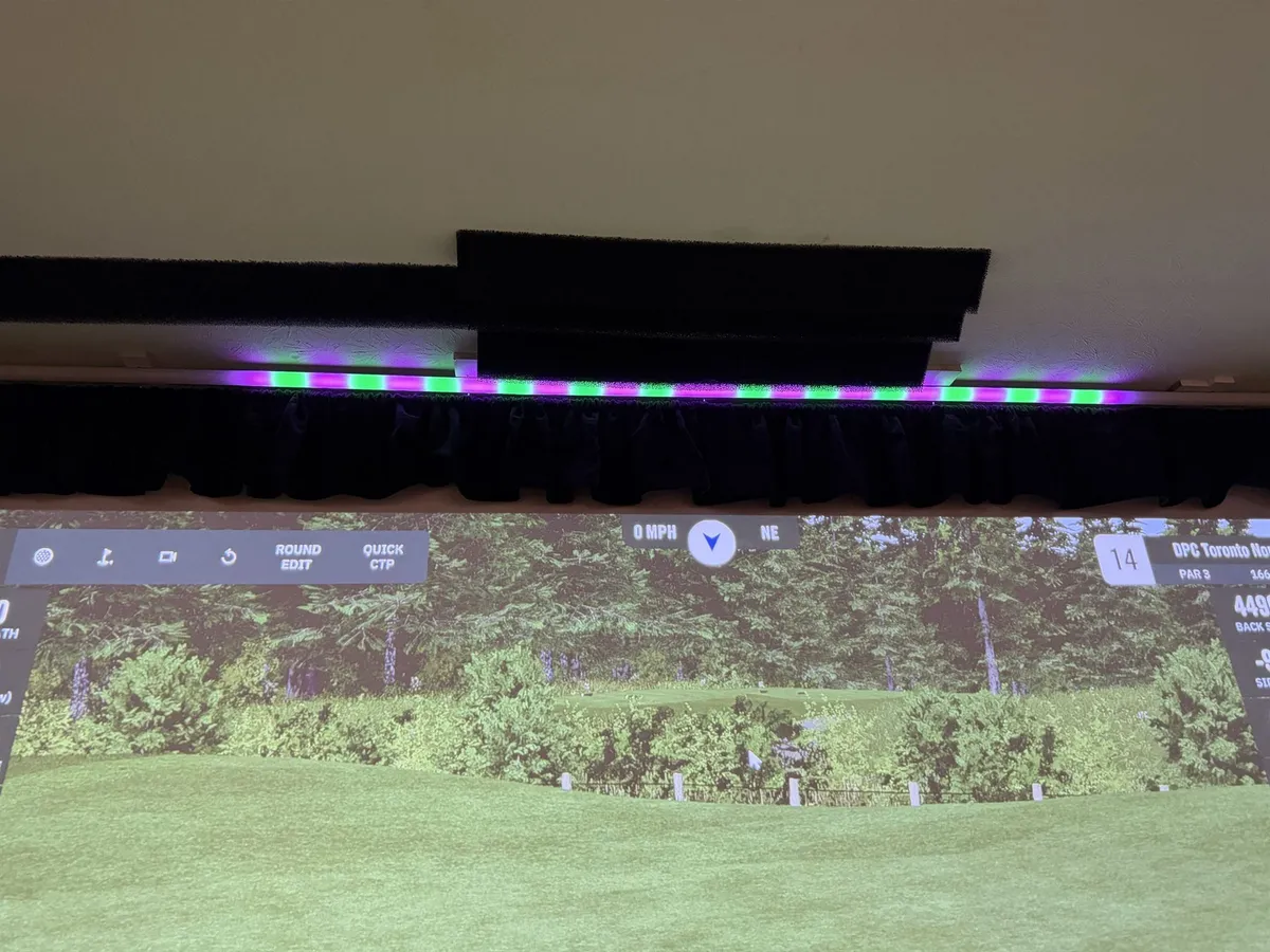 An LED light bar above a golf-sim screen glowing in a green-and-magenta sweep as a shot curves back toward the target line.