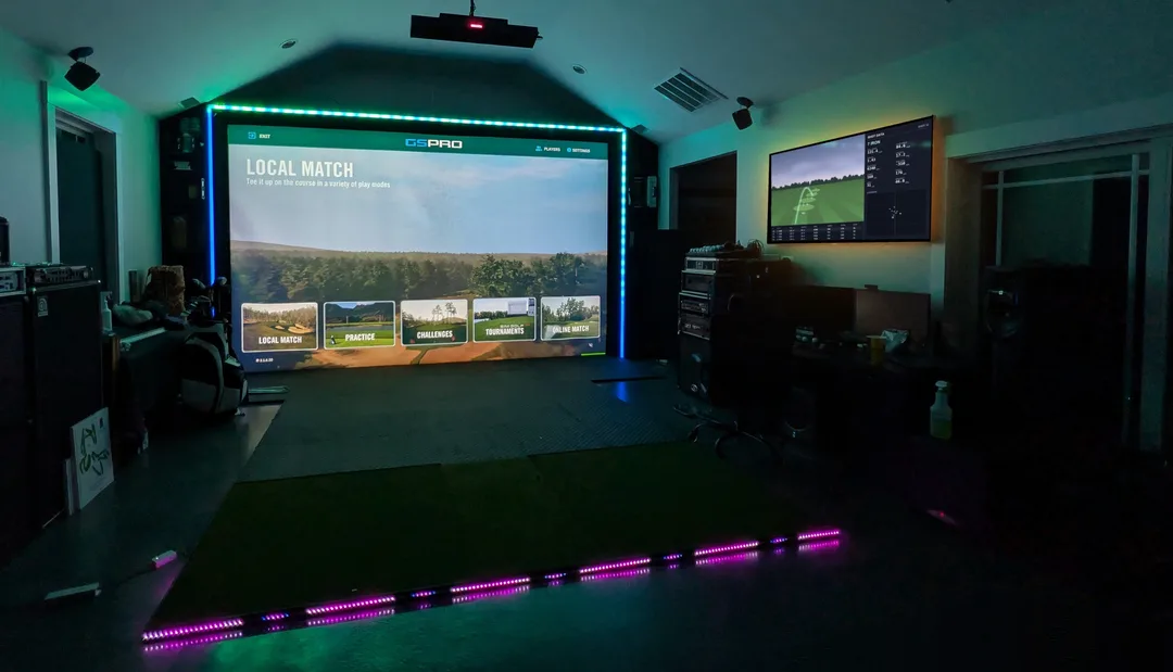 A darkened indoor golf-sim bay. An LED strip frames the projection screen and a second strip runs along the floor, both cycling through colors with their glow filling the room. GSPro is on the screen, and a wall-mounted TV shows live shot data.