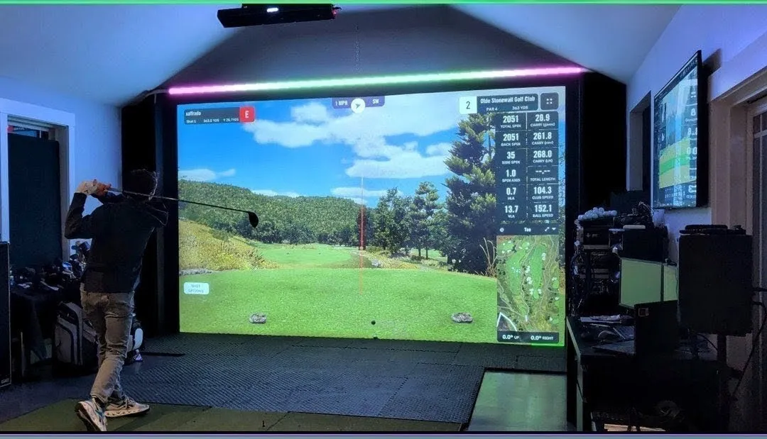 A golfer mid-swing in an indoor golf-sim room. The LED strip above the projection screen glows pink and green in reaction to the shot while GSPro shows the shot data on screen.